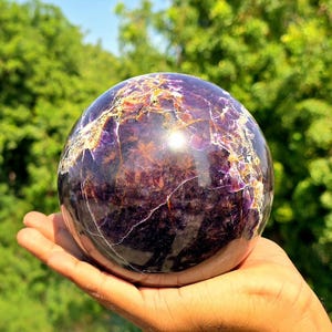 Peut inclure: Une pierre sphérique polie présentant des teintes violettes profondes avec des veines blanches et brun doré complexes sur toute la surface. La sphère affiche une texture lisse et réfléchissante avec des motifs cristallins naturels visibles.