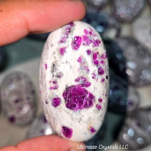 May include: An oval-shaped gemstone with a white base and vibrant pink ruby inclusions. The polished stone is held between fingers, showcasing its natural beauty. The text "Ultimate Crystals LLC" is visible.