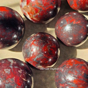 Peut inclure: Plusieurs pierres précieuses sphériques polies, présentant un mélange de motifs rouge foncé, noirs et gris. Les pierres ont une surface lisse et réfléchissante avec des variations de couleur et de veinage. Le texte "Ultimate Crystals LLC" est visible.