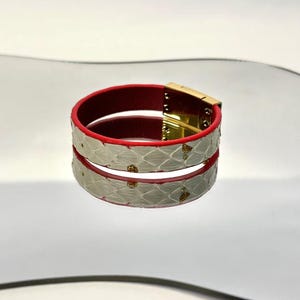 May include: A white and red leather bracelet with a gold clasp. The bracelet features a scale-like pattern on the white leather and small gold embellishments. The bracelet is approximately 2 inches in diameter.