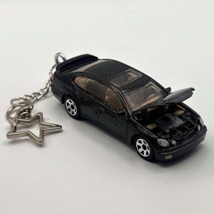 Peut inclure: Un porte-clés voiture miniature noir représentant une berline avec le capot ouvert révélant les détails du moteur, un intérieur beige, un toit ouvrant et des roues à jantes blanches. Il comprend une chaîne en métal argenté et un anneau en étoile.