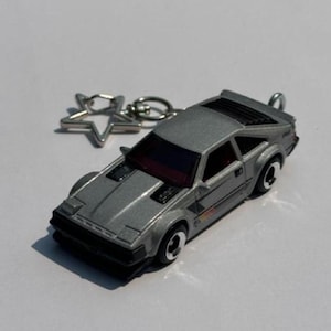 May include: A silver-colored sports car keychain featuring a metallic finish, black window detailing, and a star-shaped metal ring attachment. The miniature vehicle has a wedge-shaped body, prominent hood vents, and black wheels with white rims.