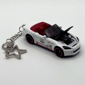 May include: A white convertible car keychain featuring a red interior, black wheels with red rims, and an open hood. The car body displays the number 333, the text S2000, and various racing decals. A silver star-shaped clasp is attached to the chain.