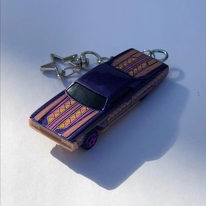 Op de afbeelding: Een paarse speelgoedautosleutelhanger met een stervormige bedel. De auto heeft een decoratief patroon in geel en roze. De wielen zijn paars. De sleutelhanger ligt op een wit oppervlak.