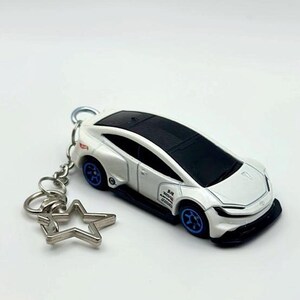 May include: A white toy car keychain featuring a black roof, blue wheels, and a silver star-shaped clasp. The car body displays small black and red graphic decals on the side panels, including the Hot Wheels logo and various text markings.