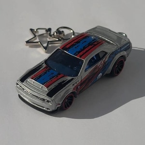 以下が含まれることがあります： 赤、青、黒のレーシングストライプが入ったシルバーのダッジチャレンジャーSRTキーホルダー。キーホルダーには星型のチャームと、赤いリムが付いた黒いホイールが付いています。車体側面には「Dodge」の文字が入っています。