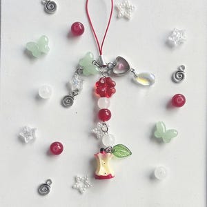 May include: A decorative charm with an apple core, red beads, and green butterfly accents. The charm is decorated with star and flower-shaped beads, and a red cord. Additional decorative beads and charms are scattered around.