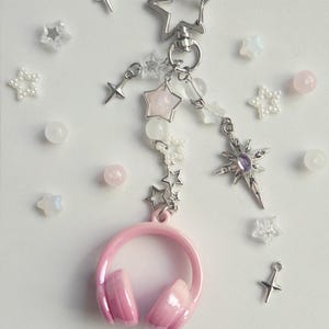 May include: A pink headphone charm with star and bead accents. The charm features a silver star-shaped clasp, a chain of small stars, and a larger star pendant. Additional star and bead embellishments are scattered around the charm.