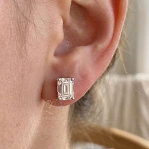 May include: A close-up of an ear with a rectangular emerald-cut diamond stud earring. The earring is set in a silver or white gold setting. The diamond has a clear, sparkling appearance.