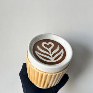 Op de afbeelding: Een keramische mok met een geribbelde, lichtbruine buitenkant en een witte rand. De bovenkant heeft een bruin cirkelvormig ontwerp met een wit hartvormig latte art-patroon in het midden, wat een realistische koffie-look op het deksel geeft.