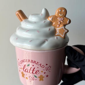 Op de afbeelding: Een lichtroze keramische mok met de tekst Gingerbread latte in rode letters, omringd door kleine peperkoekmannetjes, sterren en takjes. Het deksel is gevormd als witte slagroom met een kaneelstokje en een peperkoekmannetje.