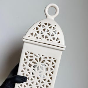 Può includere: Una lanterna in ceramica color crema con un motivo geometrico traforato su tutti i lati. La lanterna presenta una parte superiore a cupola con un'impugnatura circolare e un corpo rettangolare. Il design traforato crea un motivo a stella.
