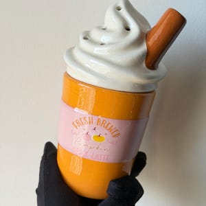 Op de afbeelding: Een keramische beker met een oranje basis en een wit deksel in de vorm van slagroom met een bruin rietje. Een roze band rond het midden toont de tekst FRESH BREWED Pumpkin SPICE LATTE met kleine illustraties van pompoenen en hartjes.
