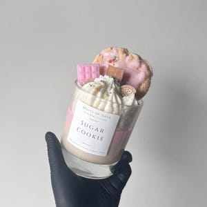 Op de afbeelding: Een glazen kaars met lagen roze en beige was, afgewerkt met witte slagroom van was, een roze koekje, een klein roze chocoladereepje en een mini sandwichkoekje. Het etiket vermeldt House of Safa Sugar Cookie 100% premium kokoswas 335g.