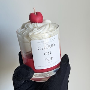 May include: A glass candle with a red base and white whipped wax topping, featuring a red cherry-shaped wax accent with a wick. The white label reads HOUSE OF SAFA, CHERRY ON TOP, and 100% natural coconut soy wax 7 OZ in black text.