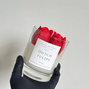 Pode incluir: Uma vela num recipiente de vidro transparente cheio de cera branca e decorado com figuras de cera em forma de tulipas vermelhas. O rótulo branco diz: HOUSE OF SAGA, Handcrafted in Amsterdam, DUTCH TULIPS, 100% premium coconut soy wax.