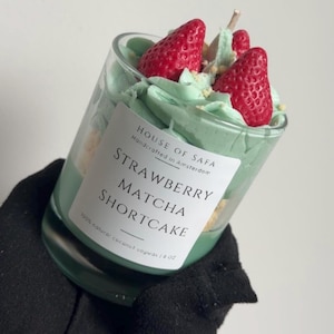 Op de afbeelding: Een glazen kaars met lichtgroene was, afgewerkt met rode aardbeivormige wasstukjes en lichtgroene opgeklopte was. Het witte etiket vermeldt: HOUSE OF SAFA, Handcrafted in Amsterdam, STRAWBERRY MATCHA SHORTCAKE, 100% natural coconut soywax.