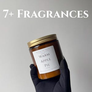 Può includere: Un barattolo per candela in vetro ambrato con coperchio dorato ed etichetta bianca con la scritta House of Safa, Warm Apple Pie e 100% natural coconut soywax 18 oz. Il testo 7+ FRAGRANCES appare sopra. La candela è tenuta da una mano con guanto nero.