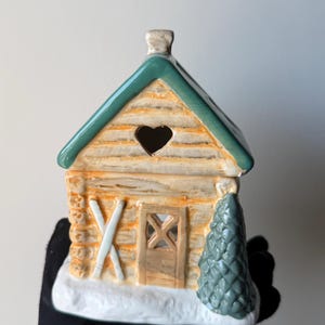Puede incluir: Una figura de cabaña de troncos de cerámica con techo verde, un recorte en forma de corazón y una base blanca nevada. La estructura incluye un exterior de color marrón claro con textura de troncos, una puerta pequeña, esquís blancos y un pino.