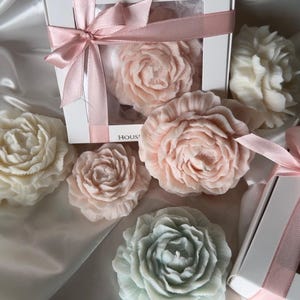 Può includere: Saponi a forma di fiore nei toni del rosa, bianco e verde. Alcuni sono in una scatola bianca legata con un nastro rosa. I saponi sono disposti su una superficie di raso. I saponi sono progettati per assomigliare a rose e peonie.