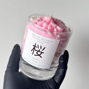 Könnte beinhalten: Eine Kerze im Klarglas mit rosa Wachs in Form von Kirschblütenblättern. Das weiße Etikett trägt die Aufschrift HOUSE OF SAFA, Handcrafted in Amsterdam, das japanische Kanji für Kirschblüte, SAKURA, und 100% natural coconut soywax 482 g.