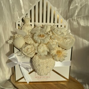 May include: A white envelope-shaped box with gold trim containing an arrangement of white wax flower-shaped candles and a small dress figurine. The box features a white satin ribbon bow and the text HOUSE OF SAFA HOME & LIVING printed on the front.