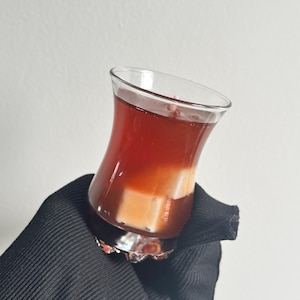 Pode incluir: Um recipiente de vidro transparente com silhueta curva contendo um líquido castanho-avermelhado escuro e dois insertos de cera brancos em forma de cubo. O vidro tem uma borda alargada e uma base espessa e texturizada com aspeto estratificado.