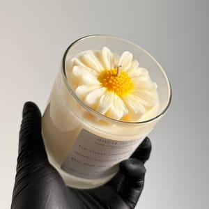 Può includere: Una candela in cera bianco sporco in un barattolo di vetro trasparente, sormontata da un fiore di margherita scolpito bianco e giallo. L'etichetta riporta House of Safa, Handcrafted in Ahmed, Top: Violet, Chamomile, Middle: Gardenia, Jasmine, Base: Musk, White Wood.