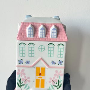 Op de afbeelding: Een keramische bus in de vorm van een huis met een witte gevel, een roze dak met drie dakkapellen en een gele deur. De buitenkant is versierd met geschilderde groene raamkozijnen, blauwe en paarse bloemaccenten en roze bloemen.