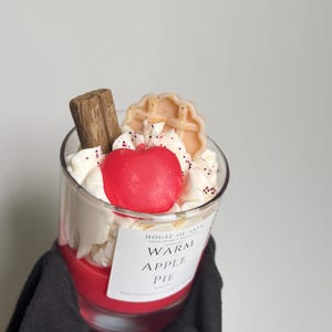 May include: A glass candle filled with red wax, topped with white whipped wax, a red apple-shaped wax piece, a brown cinnamon stick, and a beige waffle-shaped wax piece. The label reads House of Saja, Warm Apple Pie, and 100% natural.