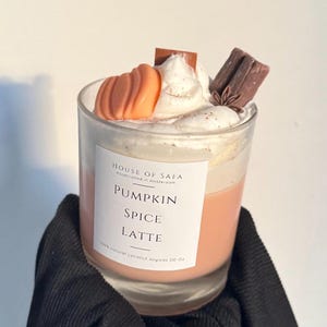 Op de afbeelding: Een glazen kaars met een wit etiket met de tekst House of Safa Pumpkin Spice Latte 10 oz. De kaars heeft een lichtoranje wasbasis, bekroond met witte slagroom van was, een kleine oranje pompoenvorm, een bruin kaneelstokje en een steranijs.