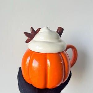 Op de afbeelding: Een feloranje keramische mok in de vorm van een pompoen met een deksel dat eruitziet als een toef witte slagroom. Het deksel is voorzien van een klein bruin kaneelstokje en een donkerbruine steranijs bovenop de witte roomlaag.
