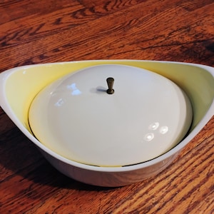 May include: A white and pale yellow ceramic serving bowl with a matching domed lid. The bowl features a wide, curved rim and an elongated oval shape. A small, dark metallic knob is centred on top of the lid for lifting. The exterior is white.