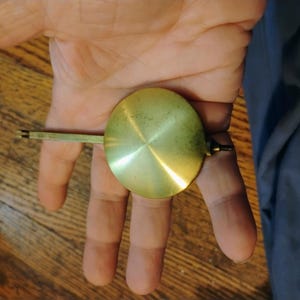 以下が含まれることがあります： 中央に点がある滑らかで反射性の高い表面を持つ円形の真鍮製時計振り子。片側から細い平らな金属棒が伸びており、時計のメカニズムに統合するための小さな切り欠きのある取り付けポイントで終わっています。