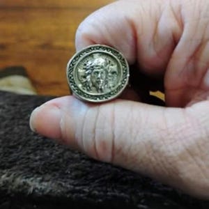 Könnte beinhalten: Ein silberfarbener Ring mit einer runden Vorderseite, die ein erhabenes, detailliertes Relief eines Gesichts mit langem Haar zeigt, umgeben von einem dekorativen Rand mit einem komplexen Muster. Das Metall ist poliert.