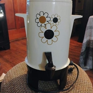 May include: A white electric beverage urn with a black base and a black spigot. The cylindrical body features a graphic print of three stylized flowers with gold, black, and white petals. It includes a white lid and two side handles.