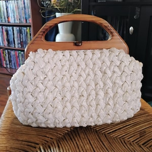 Può includere: Una borsa con corpo intrecciato bianco e un manico in legno spesso e arcuato. La borsa ha una forma rettangolare strutturata con una trama intrecciata su tutto il materiale bianco e una cornice in legno marrone lucido sull'apertura.