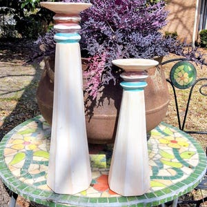 Puede incluir: Dos candelabros de madera clara, acentuados con bandas rojas y turquesas. Los candelabros se exhiben sobre una mesa con mosaicos, con una gran maceta de follaje púrpura en el fondo. Los candelabros miden aproximadamente 46 y 30 cm de altura.