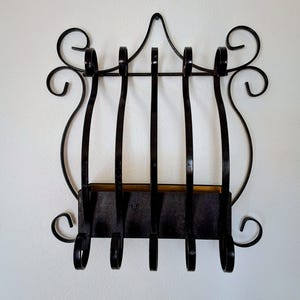 May include: A black wrought iron wall organiser with a decorative scroll design. The organiser features vertical bars and a small shelf, perfect for holding mail or small items. The design includes curved accents and hooks.