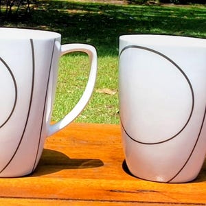 Könnte beinhalten: Vier weiße Keramikbecher mit einem schwarzen, abstrakten Design. Jeder Becher hat einen gebogenen Henkel und eine schlichte, moderne Ästhetik. Die Becher stehen auf einer Holzoberfläche, mit einem grünen, grasbewachsenen Hintergrund.
