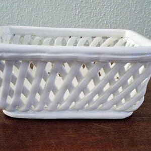 May include: A white ceramic basket with a rectangular shape and a lattice design. The basket has a smooth, glossy finish and is sitting on a dark brown surface. The basket is empty and appears to be new.