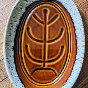 May include: Oval-shaped ceramic dish with a brown glaze and a stylized leaf design in the center. The rim has a textured, off-white and brown speckled finish. A decorative piece for serving or display.