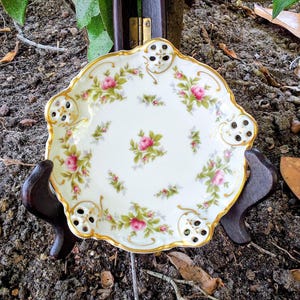 Op de afbeelding: Een witte porseleinen schaal met een geschulpte rand, versierd met roze rozen en groene bladeren. De schaal heeft een gouden rand en decoratieve uitsparingen. De schaal wordt gepresenteerd op een donkere houten standaard, met een vintage bloemenontwerp.