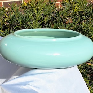 May include: A round, light turquoise ceramic bowl with a wide, shallow design. The bowl has a smooth, glossy finish and sits on a white surface. The background features green foliage and a brick wall.