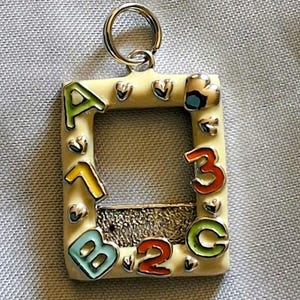 May include: A rectangular cream-colored metal charm featuring a silver jump ring at the top. The frame is decorated with colorful raised letters A, B, and C, numbers 1, 2, and 3, and small silver heart accents. A textured silver panel sits at the bottom.