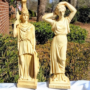 May include: Two antique statues of classical figures. One figure holds a spear and shield, wearing a helmet and draped clothing. The other figure has arms raised, with the word "TANAPRA" on the base. Both are in a light beige color.