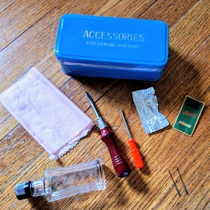以下が含まれることがあります： ACCESSORIES FOR SEWING MACHINEと書かれた青い長方形の金属製ボックス、ピンクの布、黒いキャップ付きの透明なガラス瓶、ダークレッドのハンドルのドライバー、小さなオレンジ色のドライバー、アルミホイルの小袋、緑色の針のパッケージ。