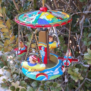 Puede incluir: Adorno de carrusel de estilo vintage con una combinación de colores rojo, amarillo y azul. El adorno presenta pequeños asientos con forma de avión con pasajeros, suspendidos de un dosel colorido. El carrusel está suspendido por un hilo dorado.