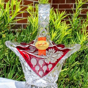 May include: A clear and ruby red lead crystal basket with a faceted handle and base. The body features etched floral patterns and a diamond-cut design. An orange sticker on the front reads Anna Hütte 24% PbO BLEIKRISTALL.