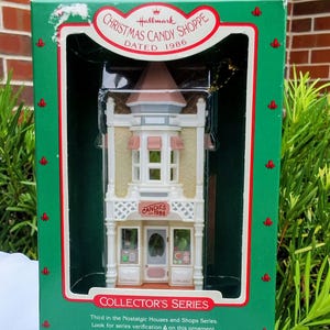 May include: A Hallmark Christmas Candy Shoppe ornament dated 1986 in its original green box. The miniature building features a cream-colored facade, a pink roof, and a storefront sign reading CANDIES 1986. The box is labeled Collector's Series.
